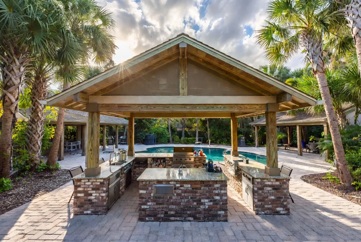 Freestanding brick outdoor kitchen under a wood pavilion beside a pool, framed by palm trees at sunset.