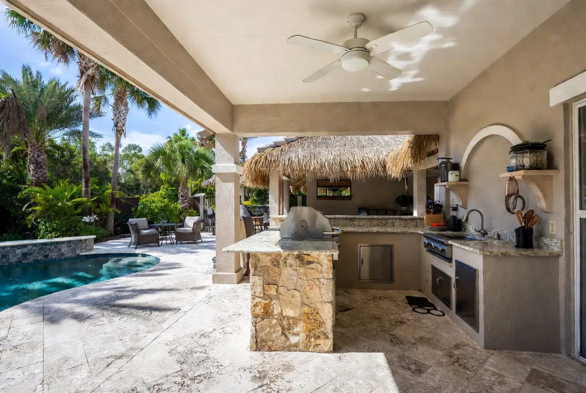 Poolside outdoor kitchen with a thatched tiki bar, stone island and spa on a travertine deck.